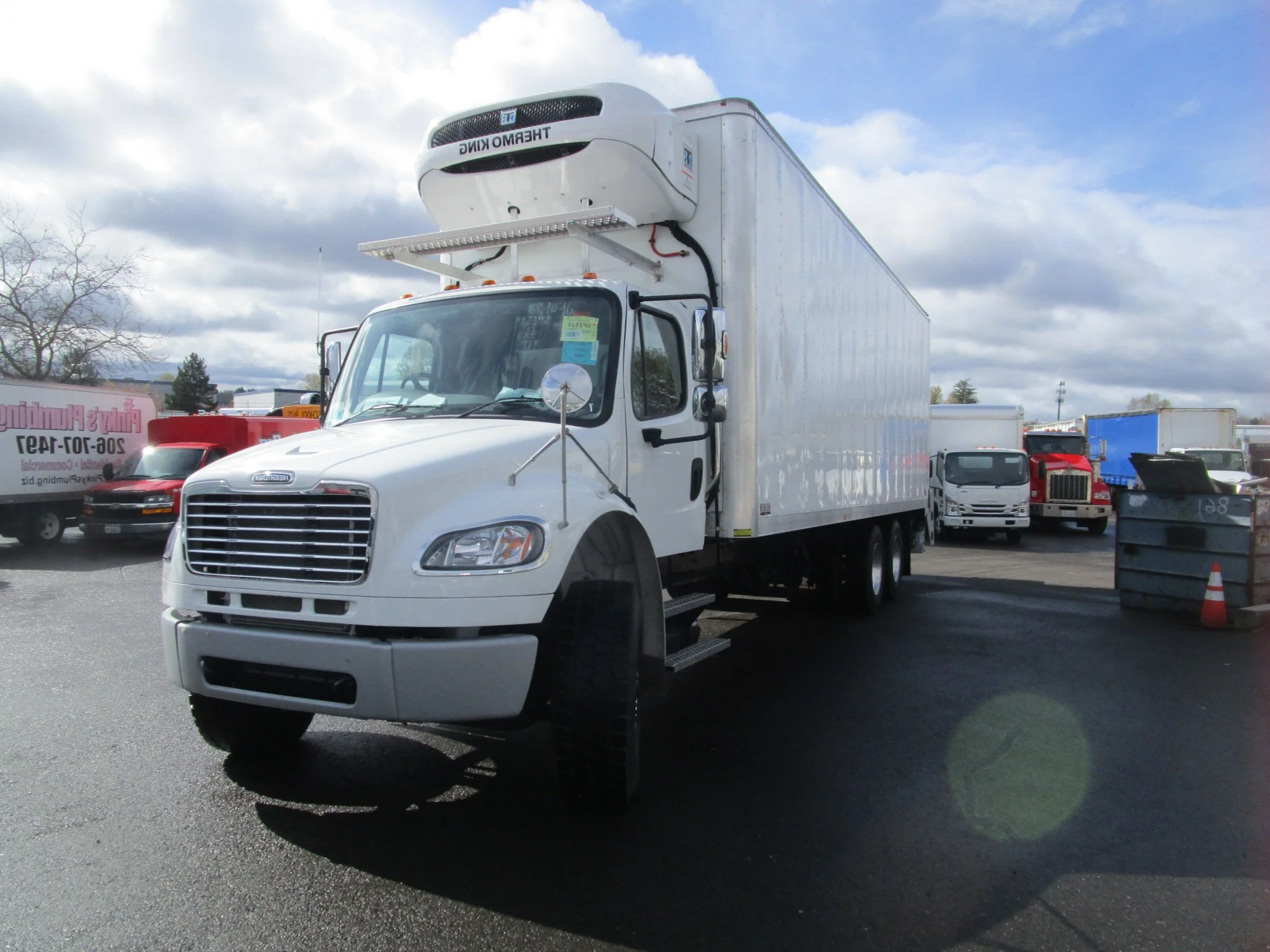 Custom installed white refrigerated box truck parked in lot with other trucks, cloudy sky above.