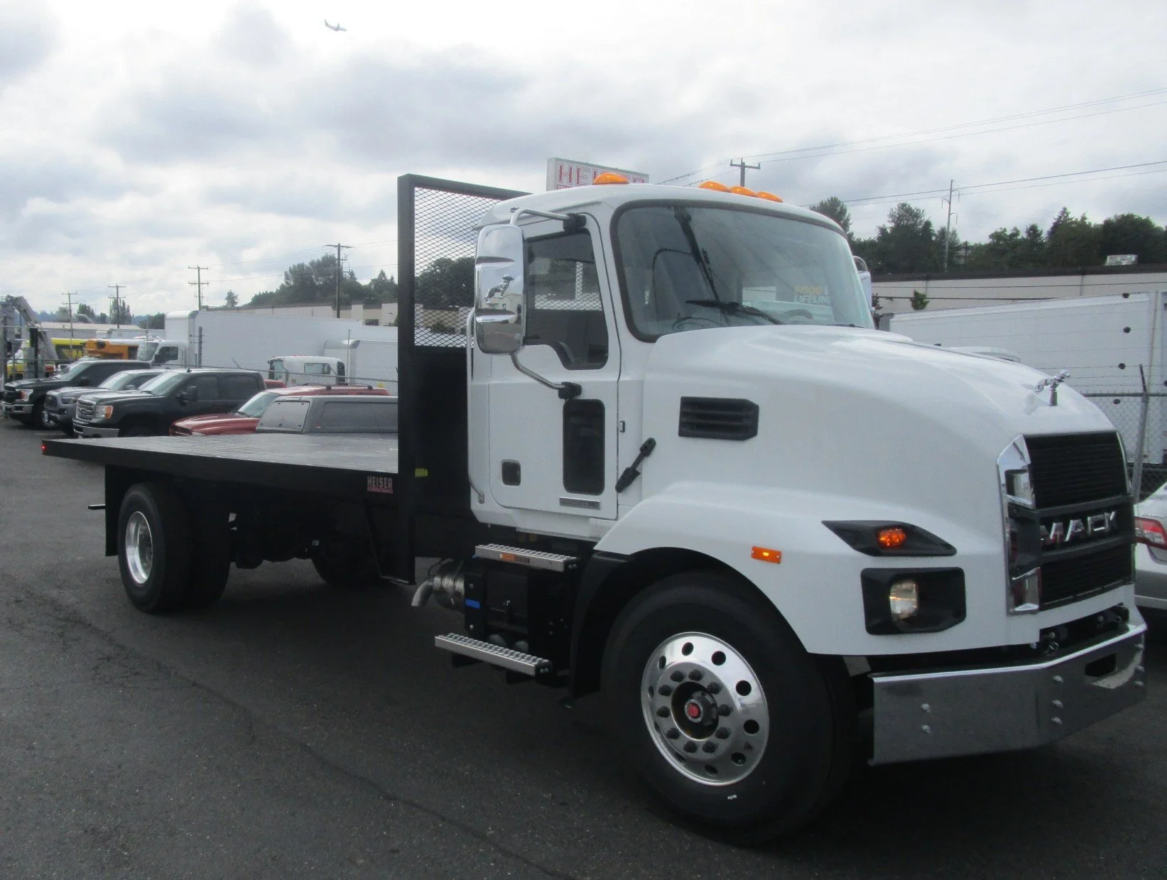 White truck with custom flatbed tow truck by Heiser Body, parked on a lot with other vehicles, cloudy sky, and power lines in the background.