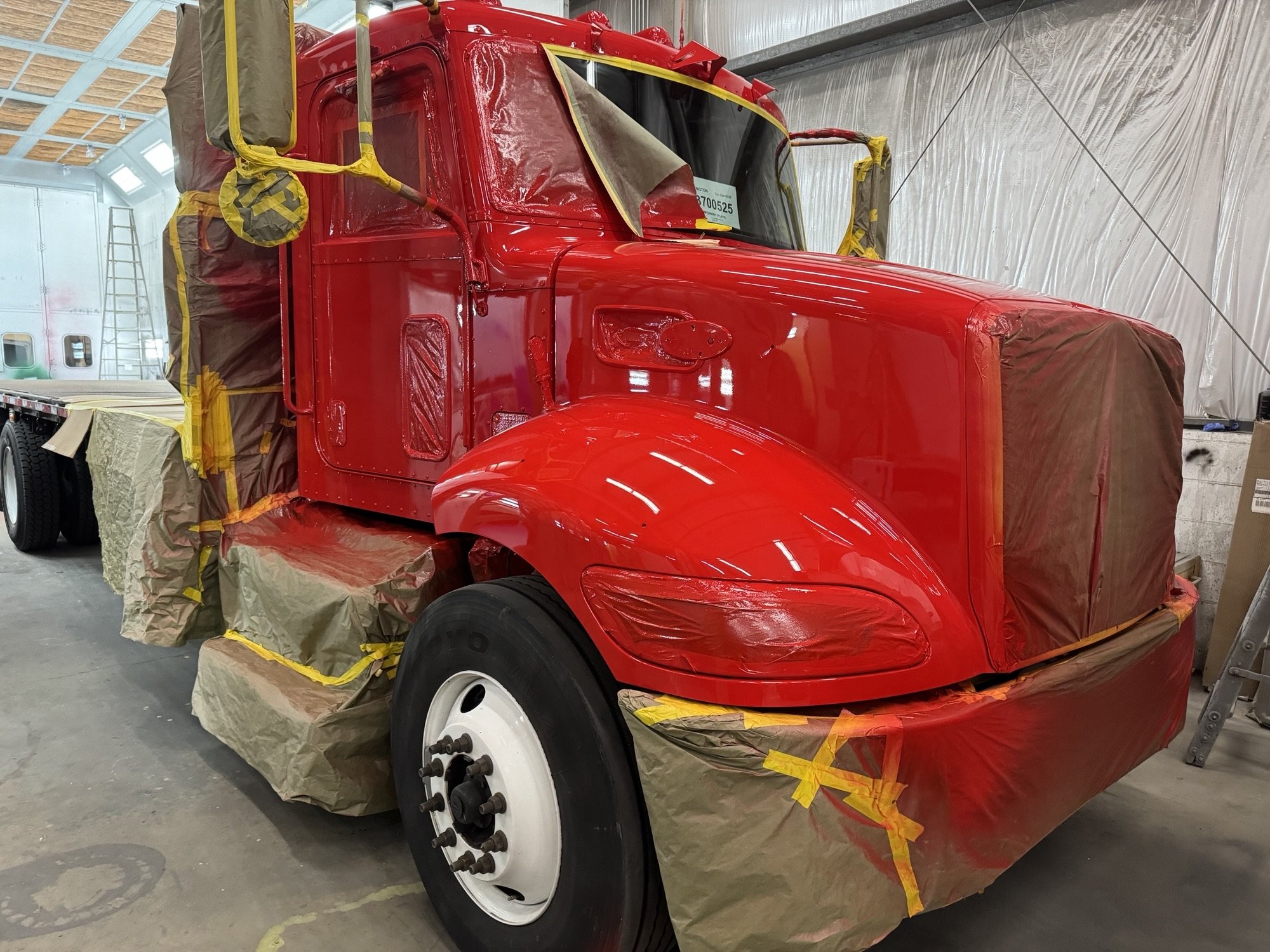 A red semi-truck cab is being painted by Heiser Body inside a workshop, with masking tape and paper covering parts of the vehicle.