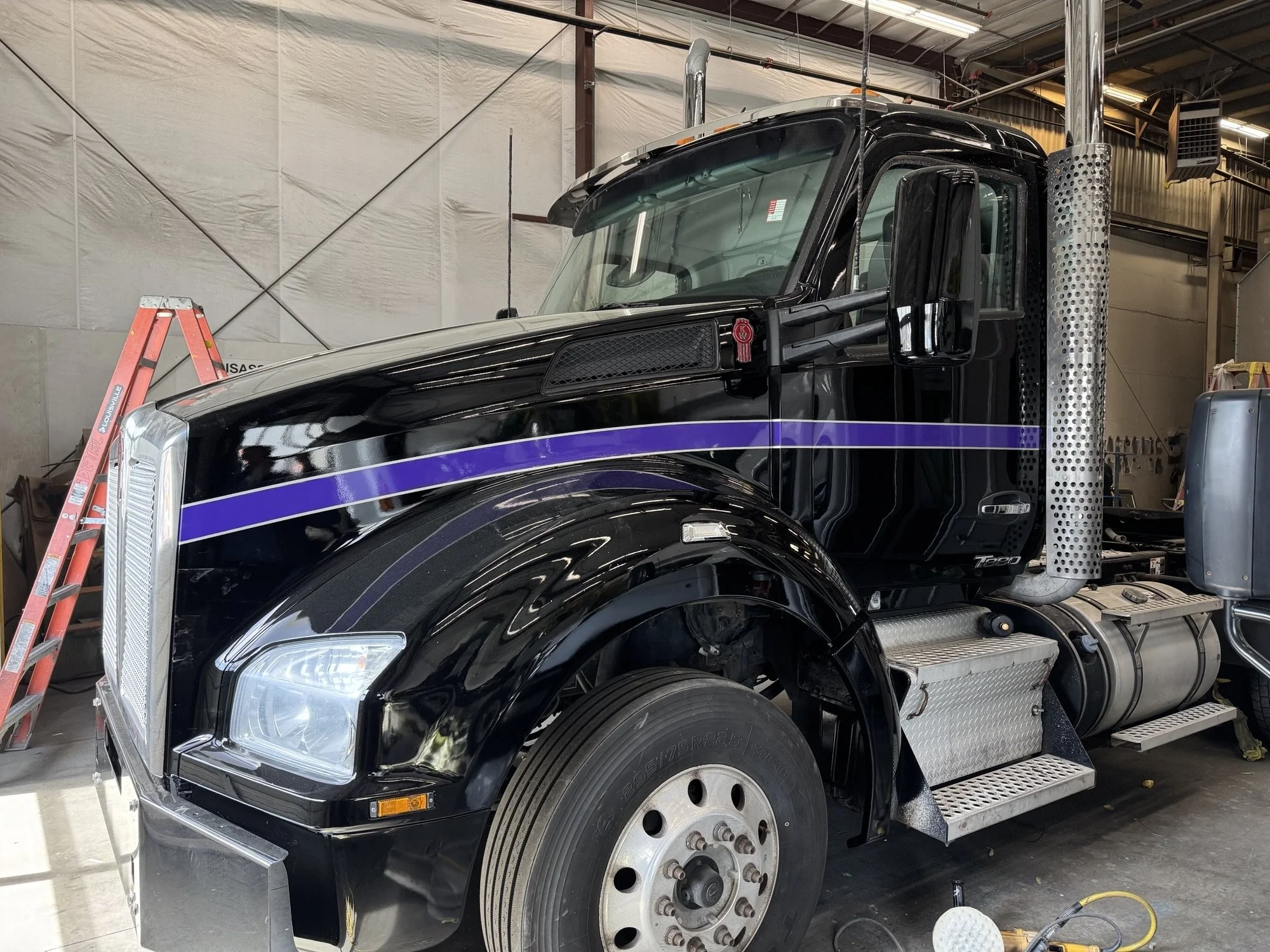 A freshly painted black semi-truck with purple stripe in a garage, with a red ladder and tools around.