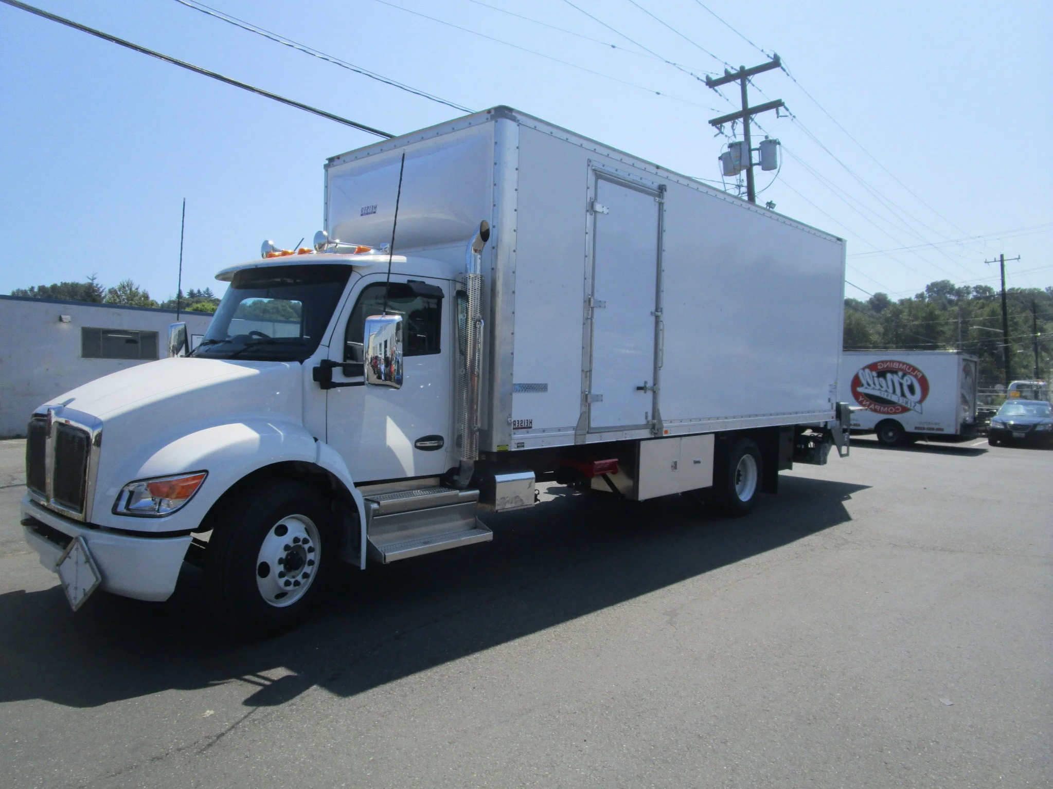 A custom installed white box truck parked on a paved lot with utility poles and other vehicles in the background.
