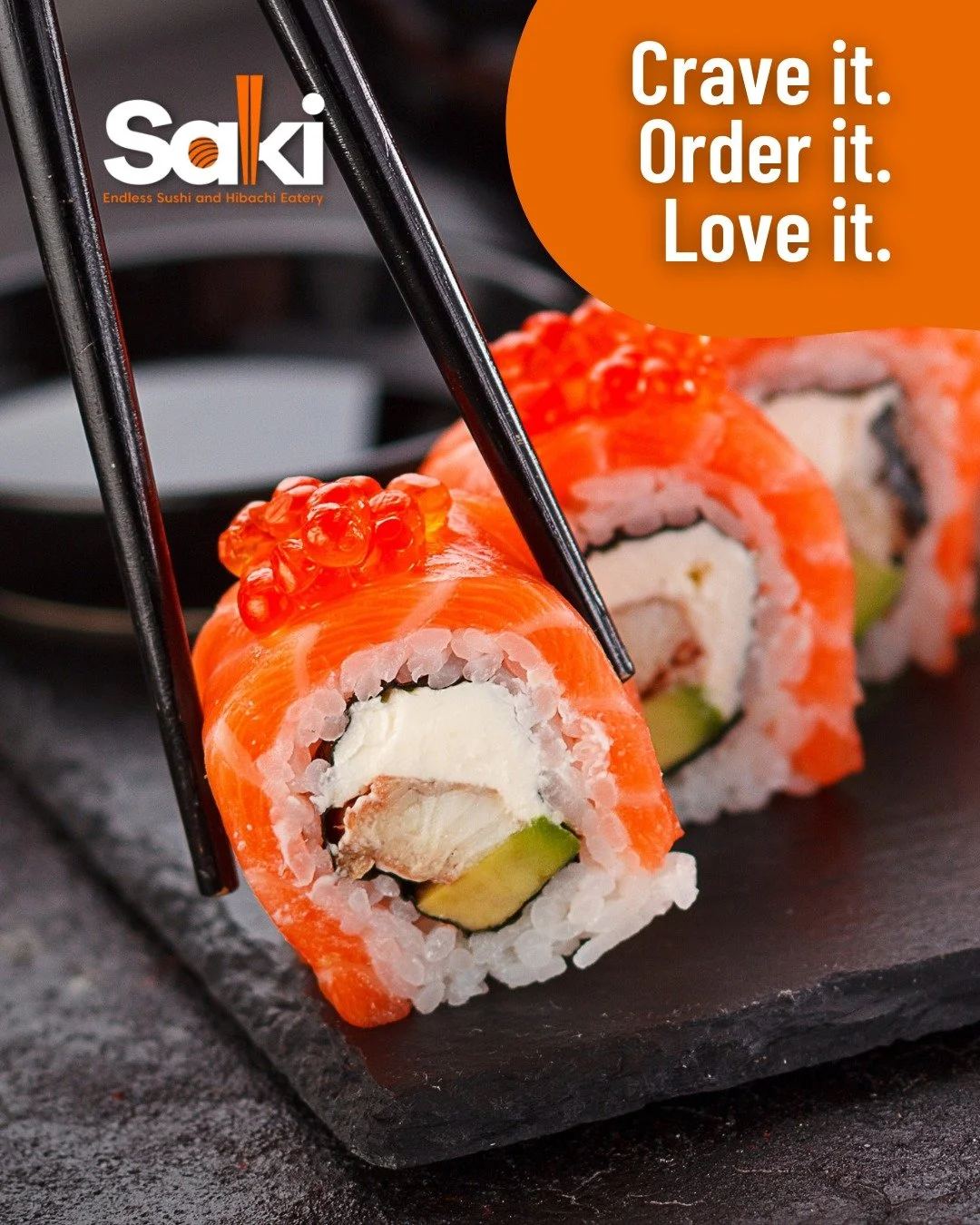 Close-up of sushi rolls with salmon, cream cheese, and avocado on a black slate serving platter. Black chopsticks lift one piece, and there is a small black dish in the background.
