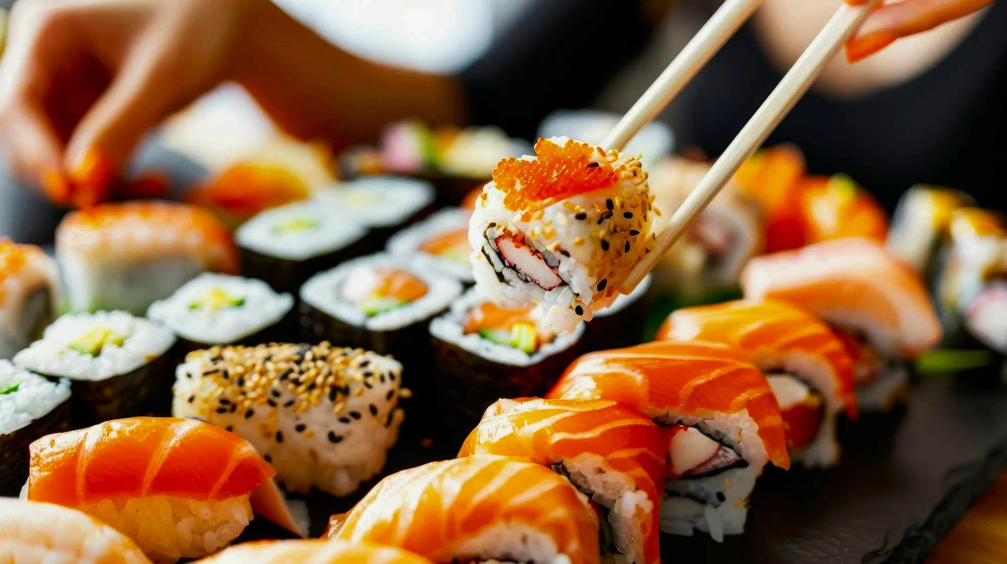 Various sushi rolls on a black platter, with chopsticks picking up one piece.