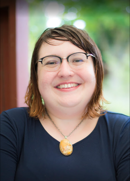 Edwina Frinefrock: a woman with short brown hair, glasses, and a necklace with a yellow pendant, smiling at the camera outdoors.
