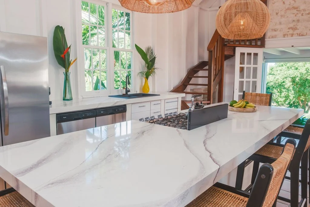 Modern kitchen with white marble island, stainless steel refrigerator, white cabinets, black dishwasher, gas stove, window with green outdoor view, wooden staircase, wicker light fixtures, and a bowl of bananas on the island.