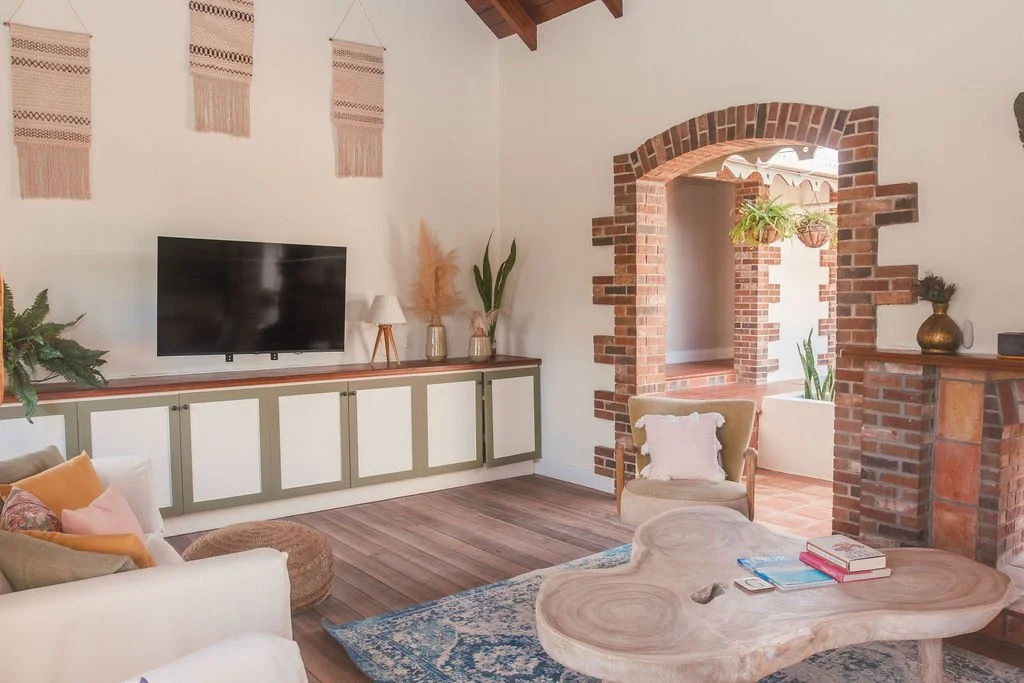Living room with brick archway, white walls, wooden ceiling beams, TV, and beige furniture with colorful pillows.