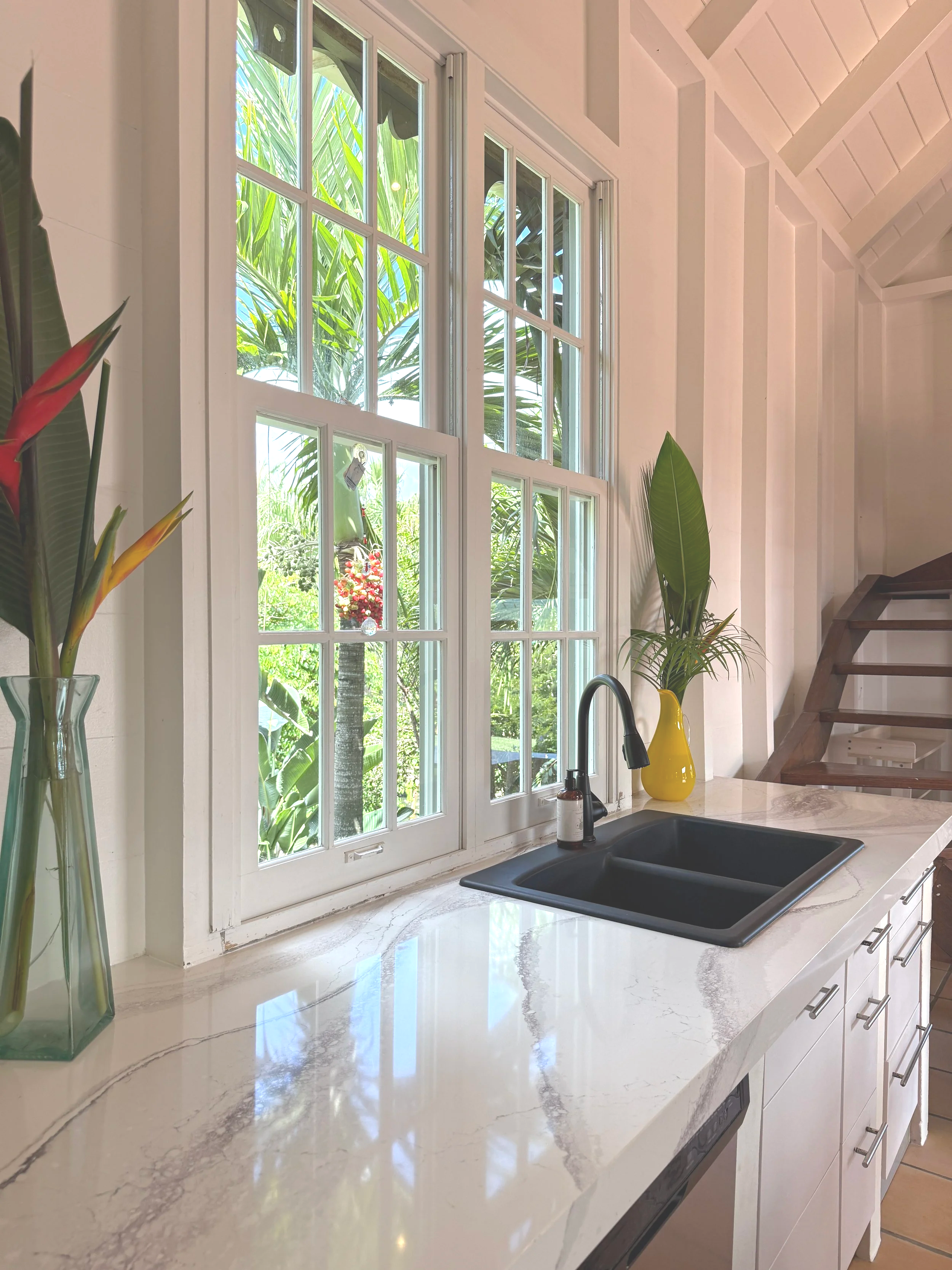 A kitchen window with white framing overlooks a lush green tropical garden. The kitchen features a white marble countertop, a black sink, and a yellow vase with a tall green plant. There are decorative items and wooden stairs visible in the backgroun