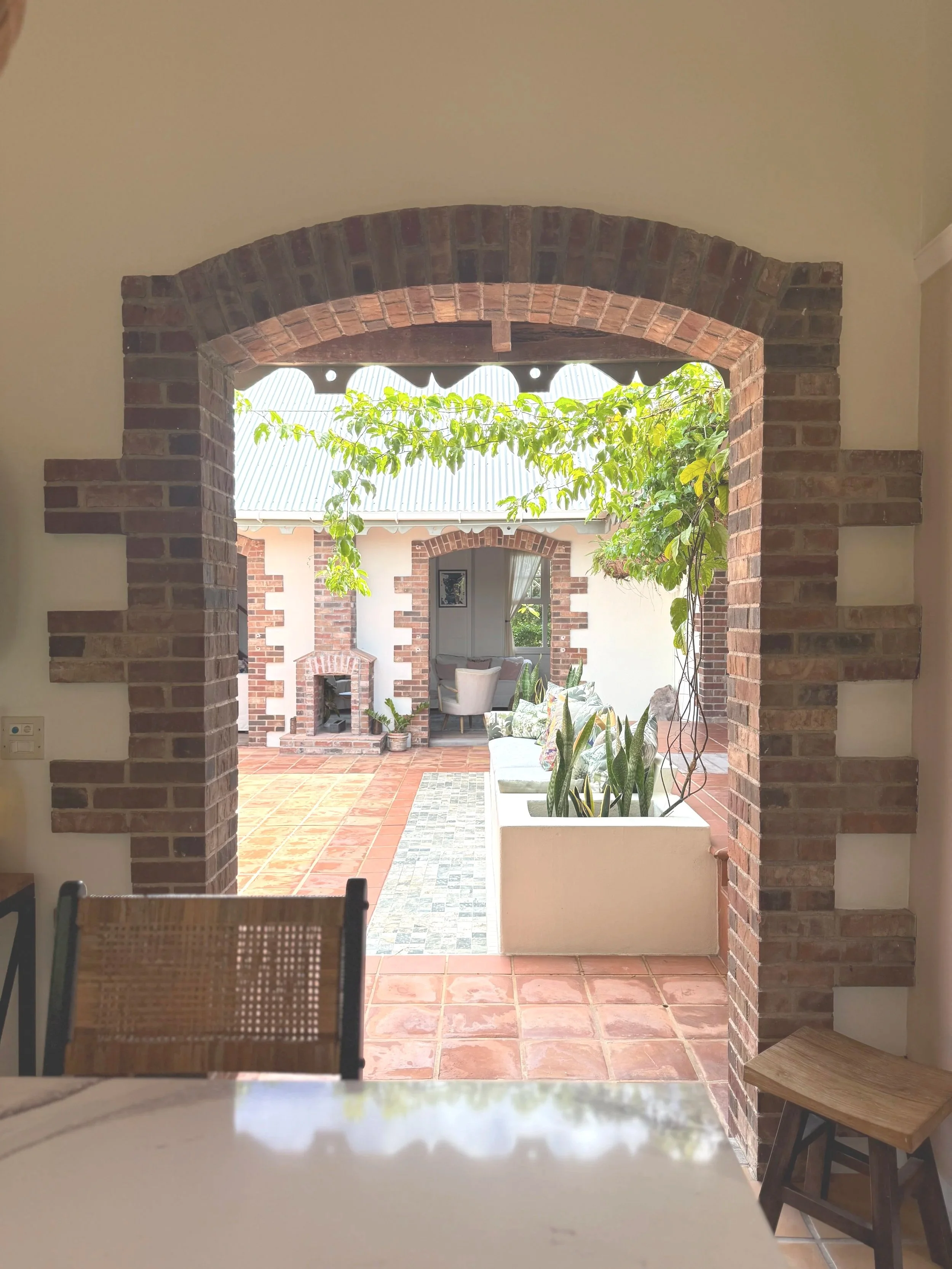 View through a brick archway into a patio with plants, a seating area, and a fireplace.
