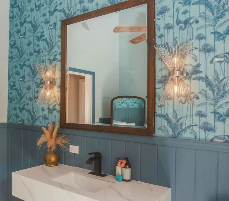 A bathroom with a white vanity, black faucet, and wall-mounted lighting fixtures. The wall has blue and green wallpaper with a tropical pattern, and a large mirror reflects parts of the room including a doorway, ceiling fan, and a bed frame.