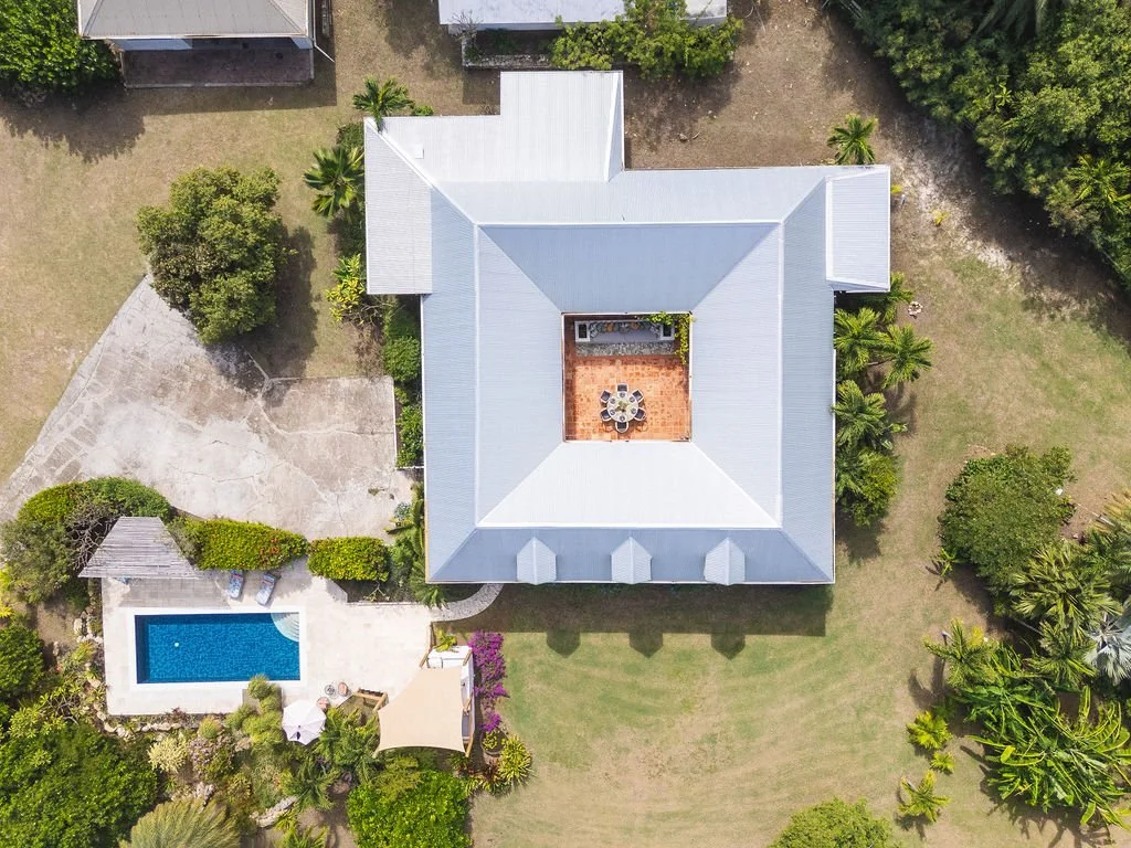 Aerial view of a house with a gray metal roof, a central patio with outdoor furniture, a backyard with a swimming pool, and lush green trees and grass.