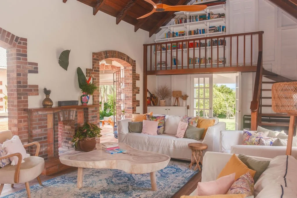 Bright living room with white sofas, colorful cushions, wooden coffee table, brick fireplace, bookshelves on loft, and large windows showing green outdoor scenery.