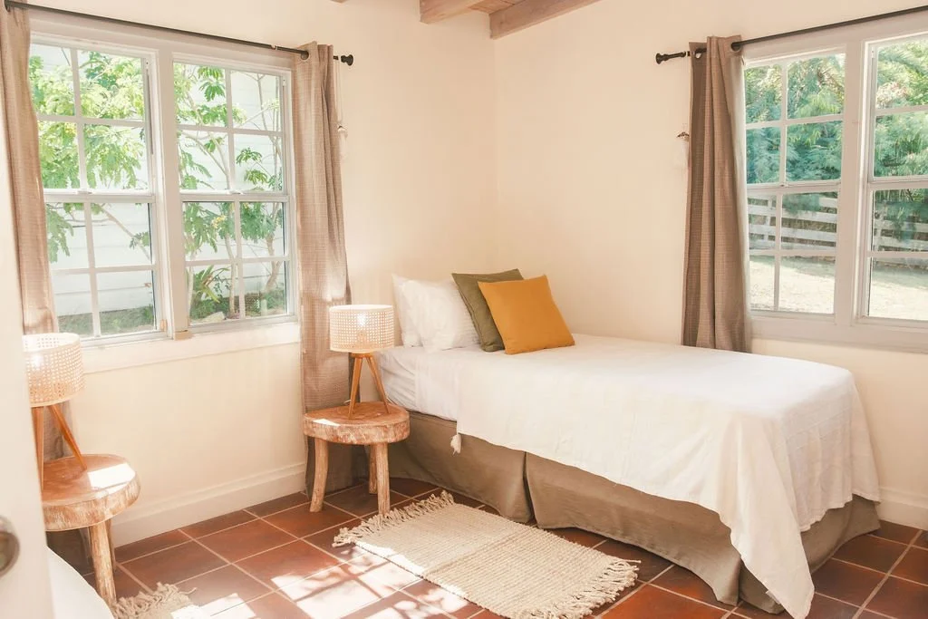 Bedroom with a bed, two windows, curtains, two wooden side tables with lamps, and a small rug on the floor.