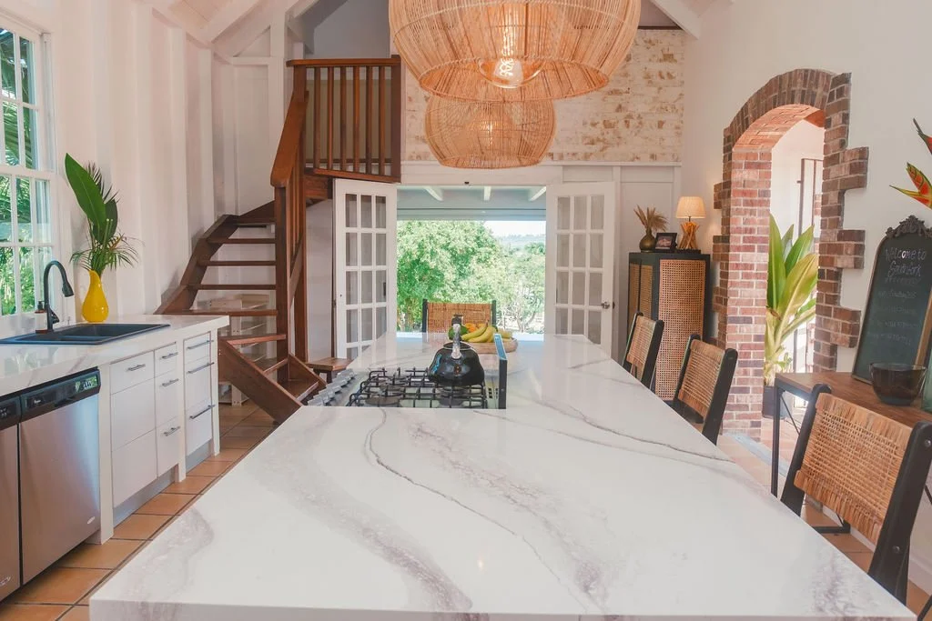 Kitchen with large white marble island, black chairs, brick archway, wooden staircase, and sliding glass doors leading to an outdoor view.