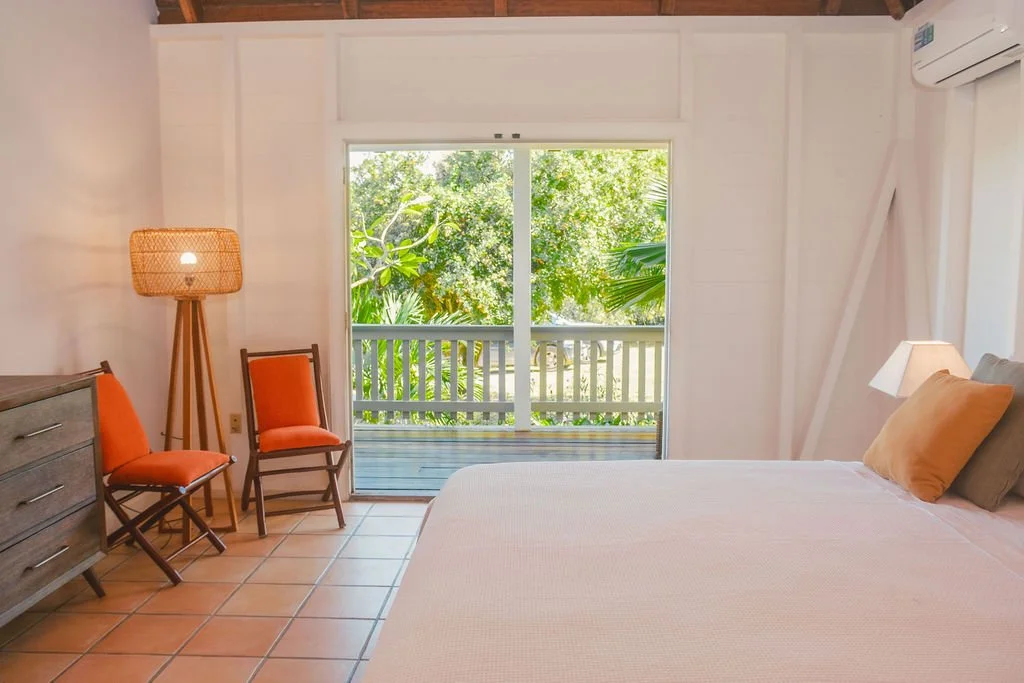 A bedroom with white walls, a large bed with beige and dark brown pillows, a nightstand with a lamp, two orange chairs, a wooden dresser, a rattan floor lamp, a sliding glass door opening to a balcony with greenery outside, and an air conditioning un