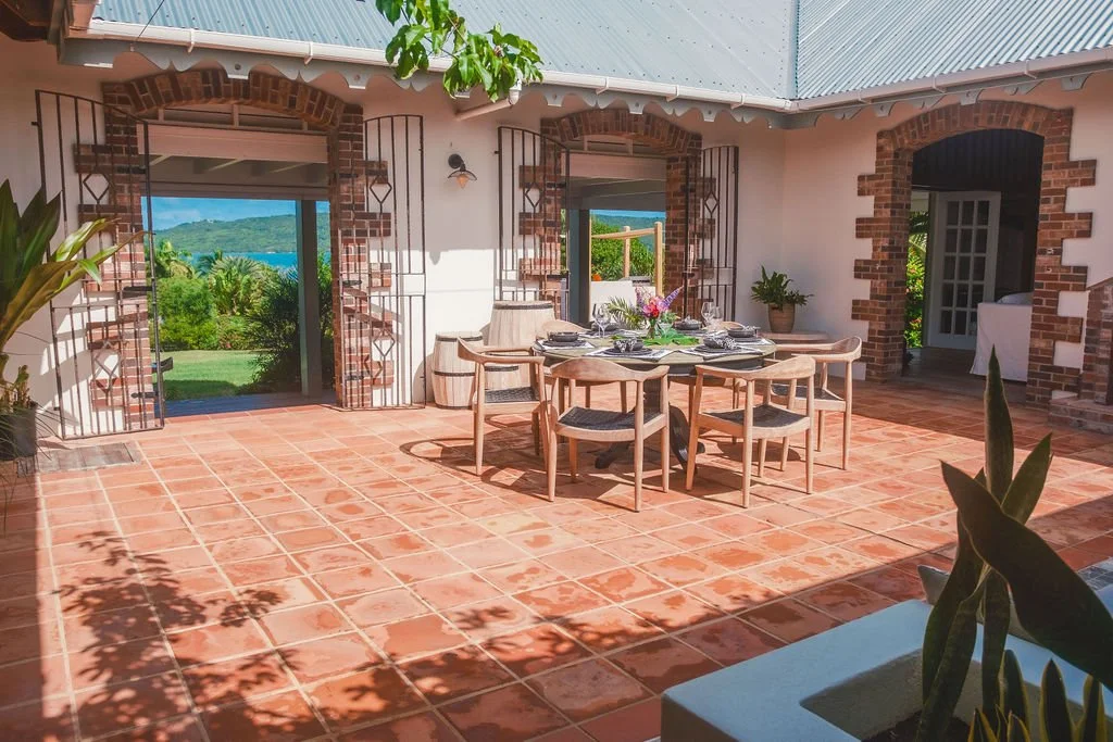 Outdoor patio with terracotta tile flooring, a round dining table set with plates, glasses, and a centerpiece, surrounded by eight wooden chairs, and a view of greenery and water through open doors.