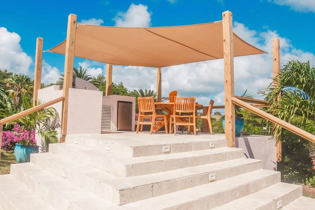 Outdoor patio with wooden dining table and chairs under a beige canopy, set on a stone platform with steps, surrounded by plants and palm trees, with a bright blue sky and clouds overhead.