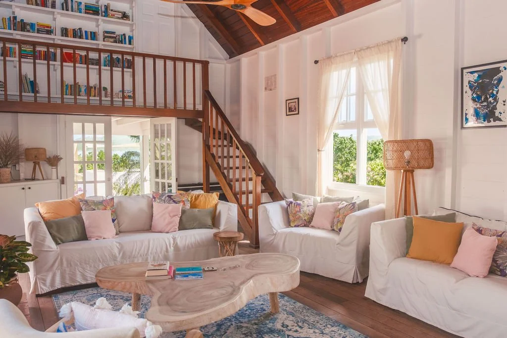 Living room with white sofas decorated with colorful pillows, a wooden coffee table, a bookshelf on the second floor, a window with curtains, a wooden staircase, and a wall art piece.