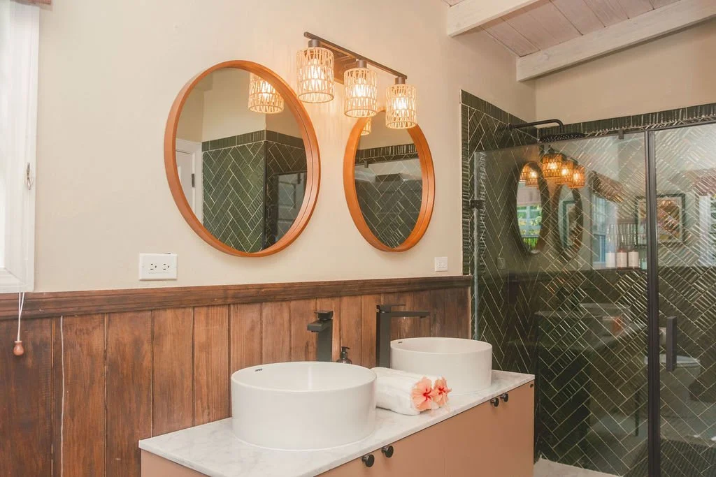A modern bathroom with two white vessel sinks on a beige vanity, wooden wall paneling, two round mirrors above the sinks, and a glass shower enclosure with black diagonal tile pattern. There are three hanging wicker light fixtures above the mirrors.
