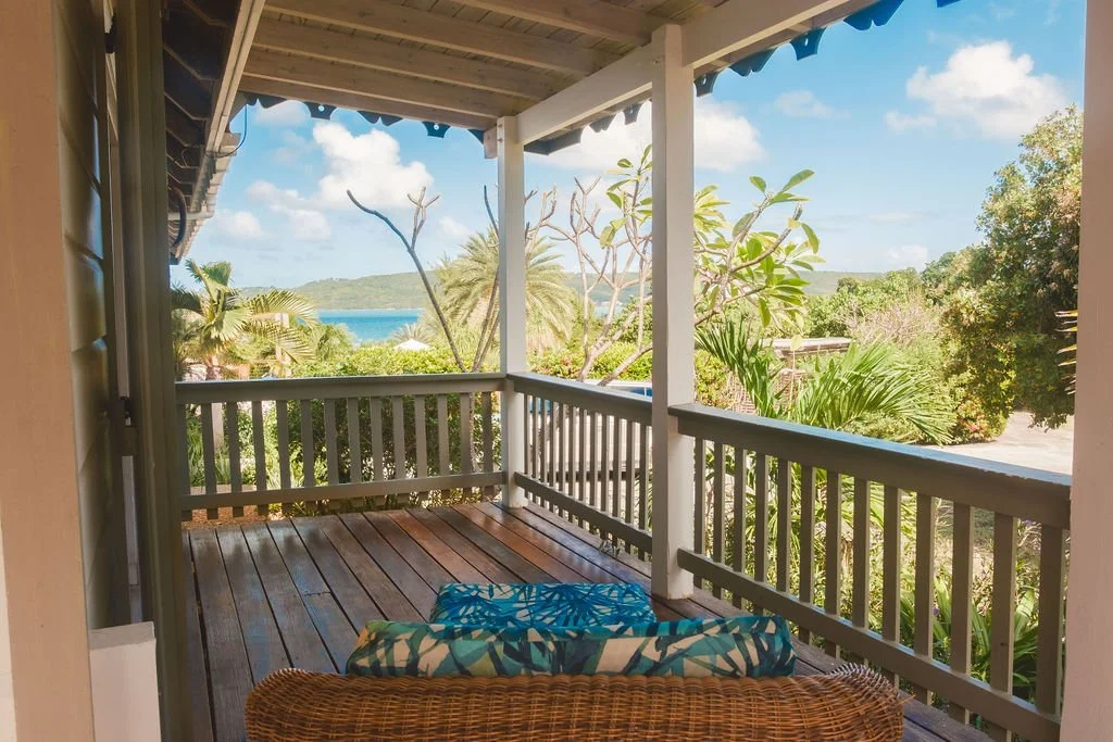 View from a porch with wooden floorboards and white railings, overlooking lush green trees, palm trees, and a glimpse of water in the distance under a partly cloudy sky.