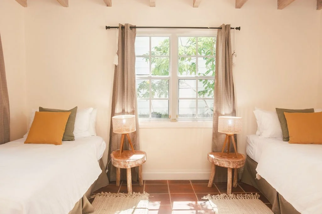 Two twin beds with white bedding and mustard yellow and gray pillows, separated by a window with beige curtains, and wooden nightstands with lamps, in a cozy bedroom with wooden ceiling beams.