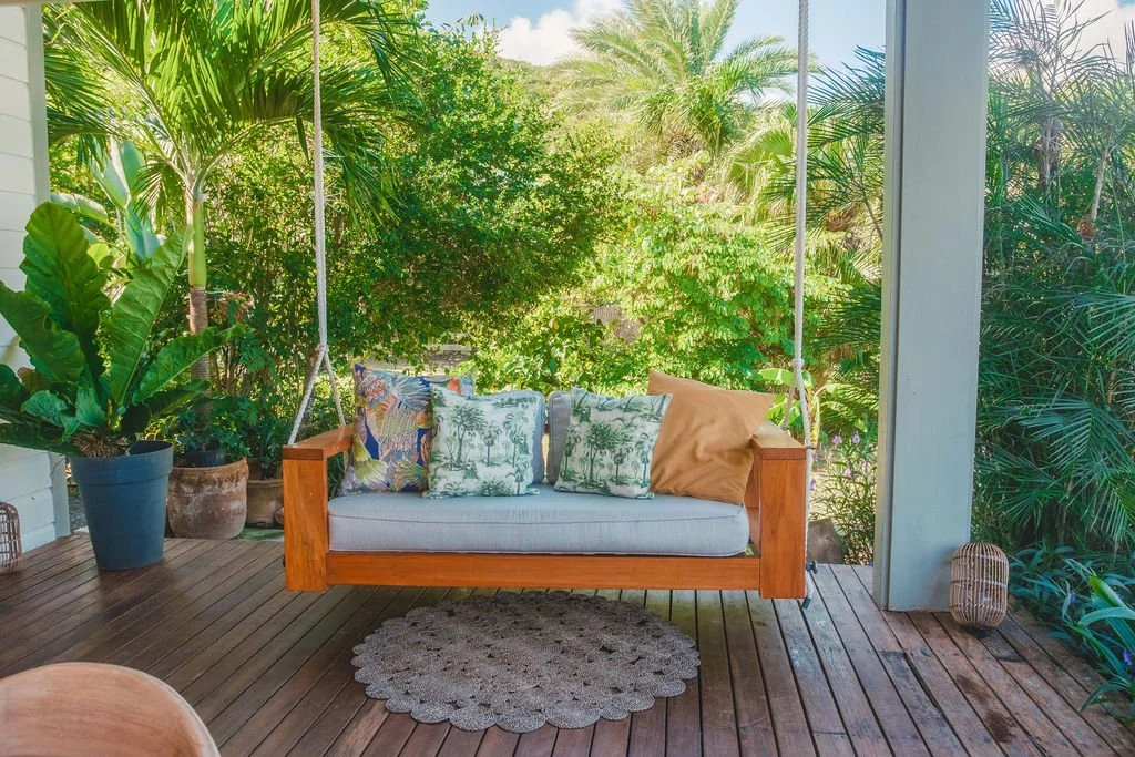 A wooden porch with a hanging bench swing surrounded by lush green tropical plants and trees, with decorative pillows on the swing.