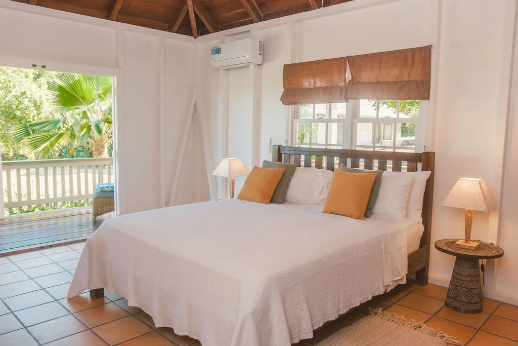 Cozy bedroom with a wooden bed frame, white bedding, and four throw pillows. Nightstand with a lamp on each side, window with bamboo shade, and a sliding door leading to a balcony with tropical plants.