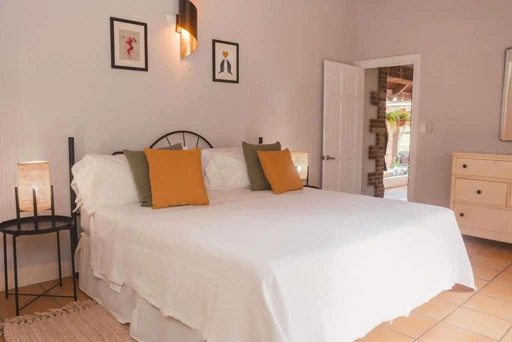 A bedroom with a large bed, white bedding, and four throw pillows in olive green and mustard yellow. There are two black bedside tables with woven lamps on each side. Artwork hangs above the bed, and a partially open door reveals part of a porch with