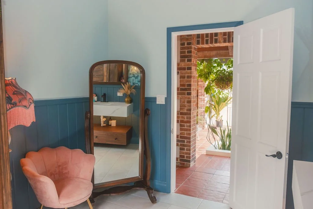 Interior view of a room with light blue upper walls and darker blue lower wainscoting, a white door partially open leading to an outdoor area with brick archways and potted plants, a pink upholstered chair, an antique freestanding mirror, and a woode