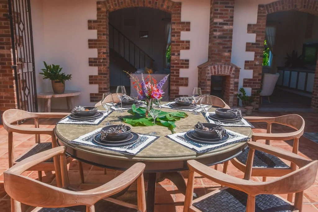 A round dining table set for six with plates, napkins, wine glasses, and a floral centerpiece in a patio area with brick walls and a fireplace.