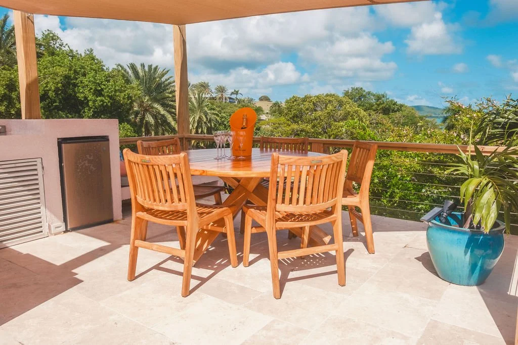 A wooden outdoor dining table with four matching chairs on a patio. A large blue planter with green leafy plants and a rolled-up yoga mat are nearby. The patio overlooks lush greenery, trees, and a partly cloudy sky.