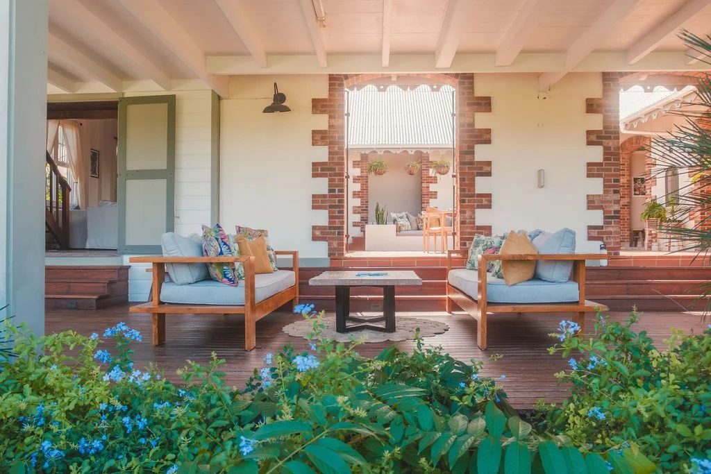 Front porch with two wooden and cushioned chairs, a small wooden table, potted plants, and a view into a cozy living room area.