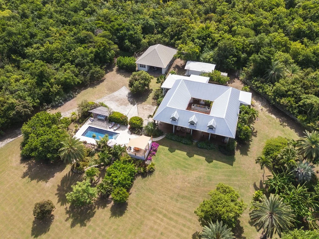 Aerial view of a large house with a metal roof surrounded by lush green trees, a swimming pool with lounge chairs and umbrellas, a comfortable patio, and open grassy areas.