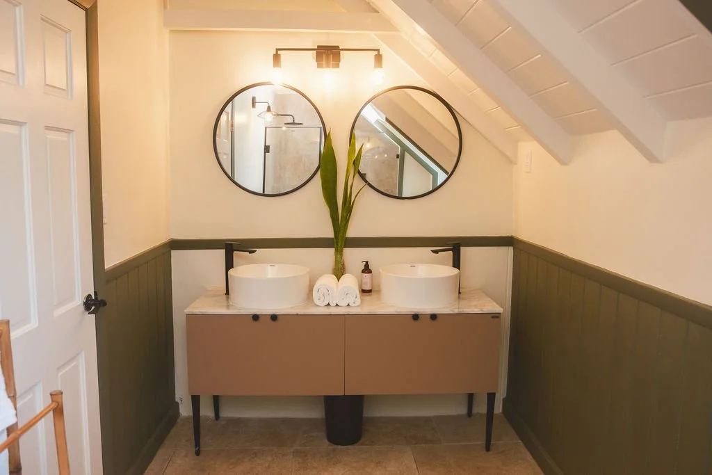 Bathroom with double vessel sinks on a vanity, two round mirrors above, a tall plant placed between the mirrors, rolled towels, soap dispenser, and a light fixture with three bulbs, all under a sloped ceiling.