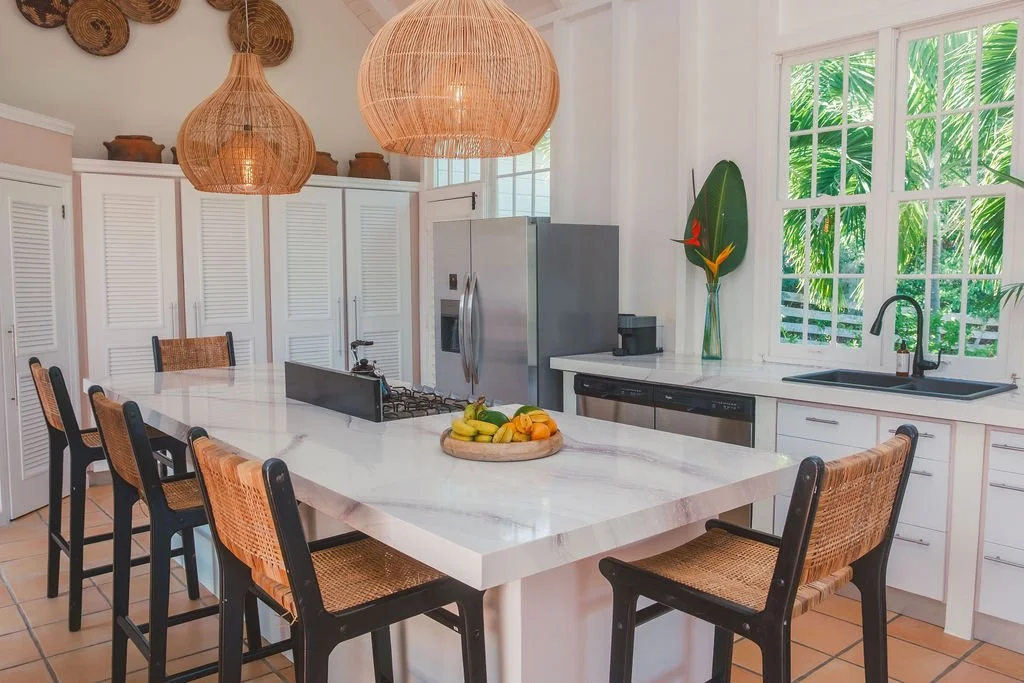Bright kitchen with white cabinets, large island with fruit bowl, stainless steel fridge, black sink, and wooden chairs; large windows with greenery outside; two large woven pendant lights hanging from the ceiling.