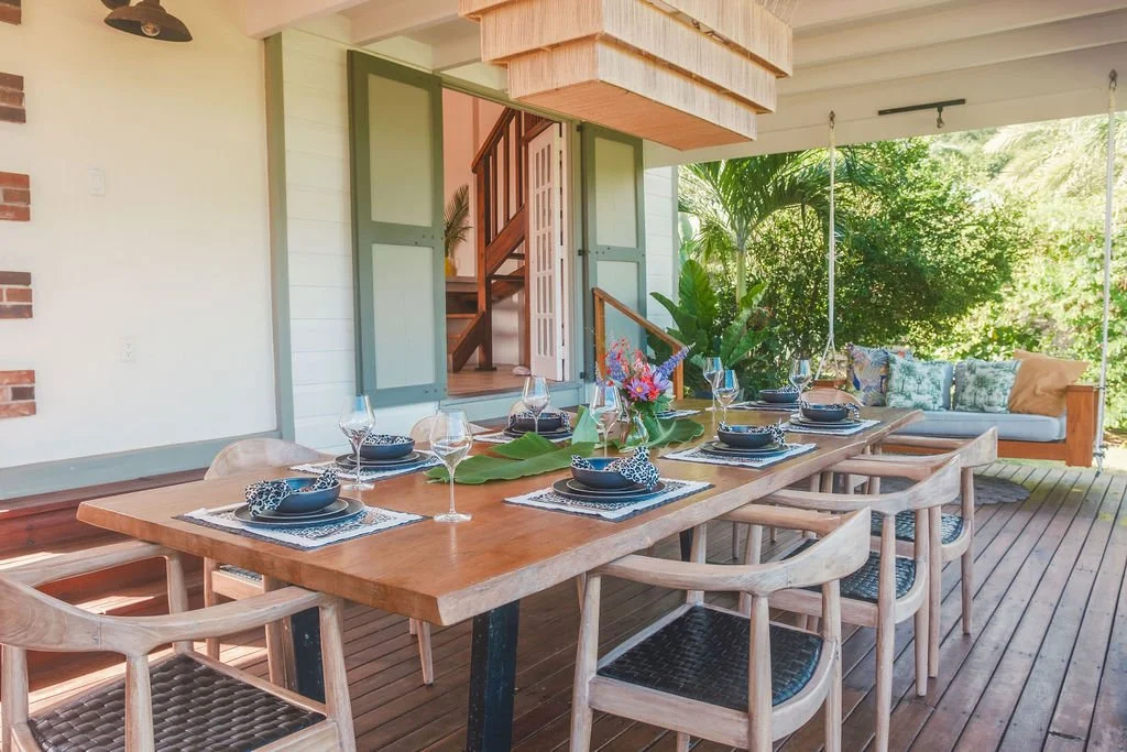 Outdoor dining area with a wooden table set for six, surrounded by matching chairs, with a floral centerpiece and black dishware. In the background, there's a garden with lush green foliage, a cushioned swing, and an open door revealing an interior s