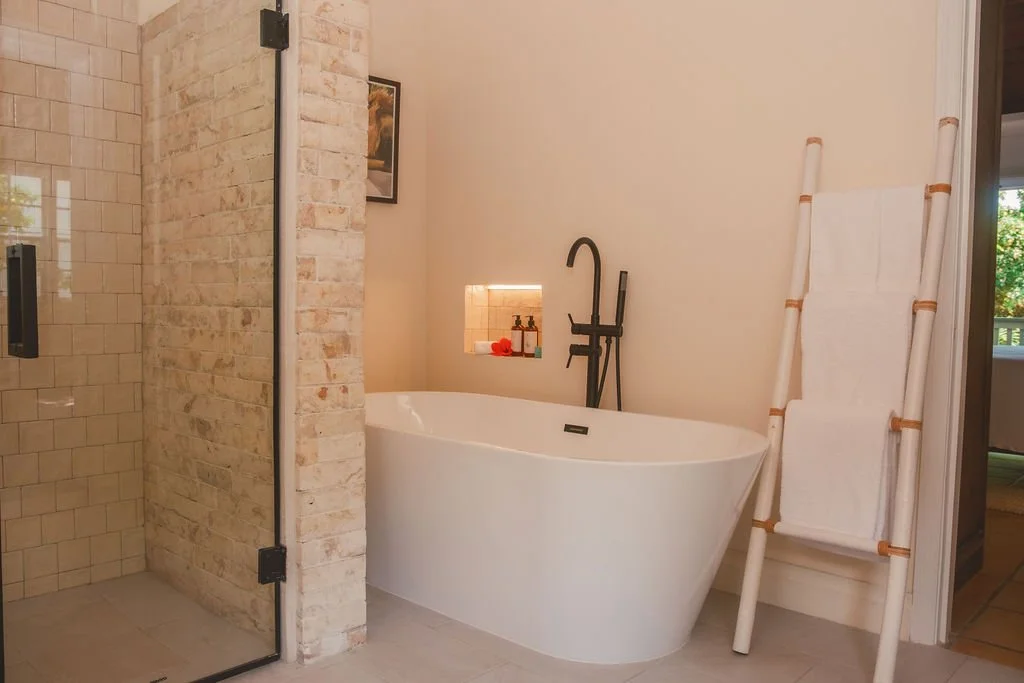 Modern bathroom with freestanding white bathtub, black faucet, and beige tile shower enclosed with glass door. White ladder towel rack and a small recessed shelf with bath products and flowers, placed against a light-colored wall.
