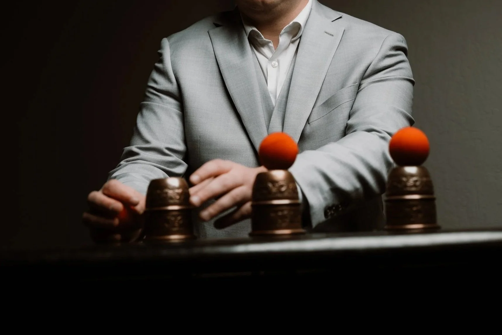 A man in a gray suit with a white shirt standing behind a table with four stacked copper cups and orange balls on top in a dark room.