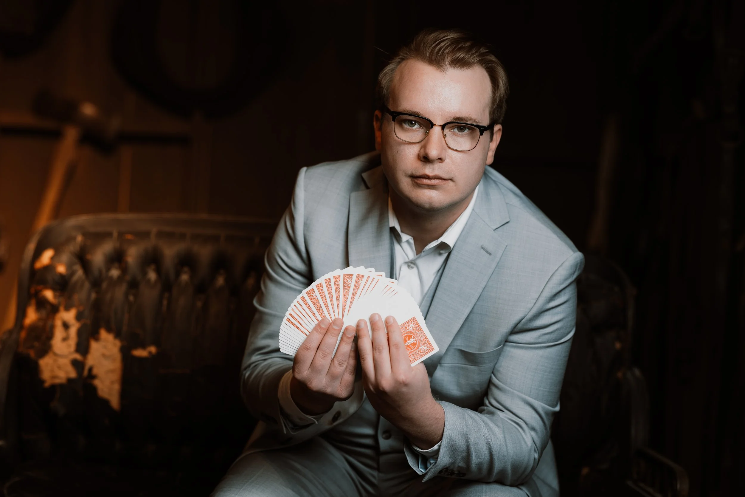 A man in a light gray suit holding a fan of playing cards, sitting on a vintage-style sofa in a dimly lit room.