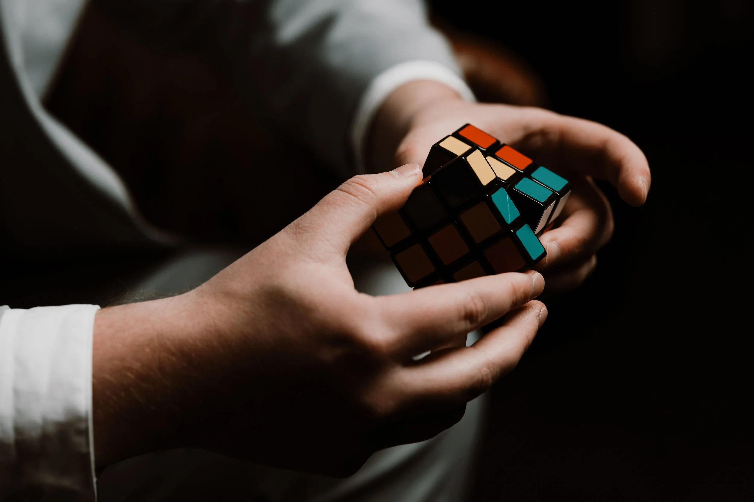 Person holding a partially solved Rubik's Cube with black edges and multicolored stickers, including orange, beige, blue, and brown, in a dimly lit environment.