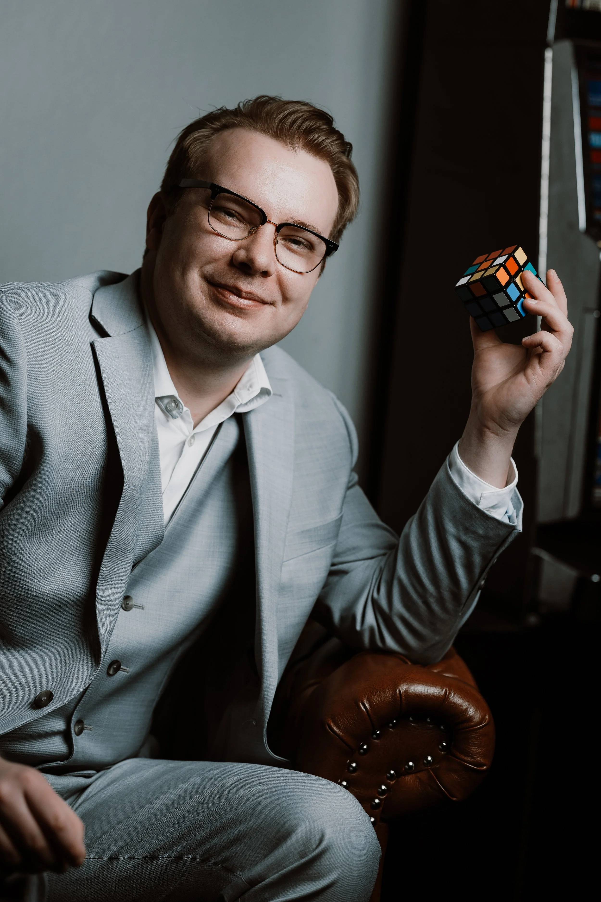 A man in a light gray suit and glasses sitting on a brown leather chair, holding a Rubik's Cube, smiling at the camera.