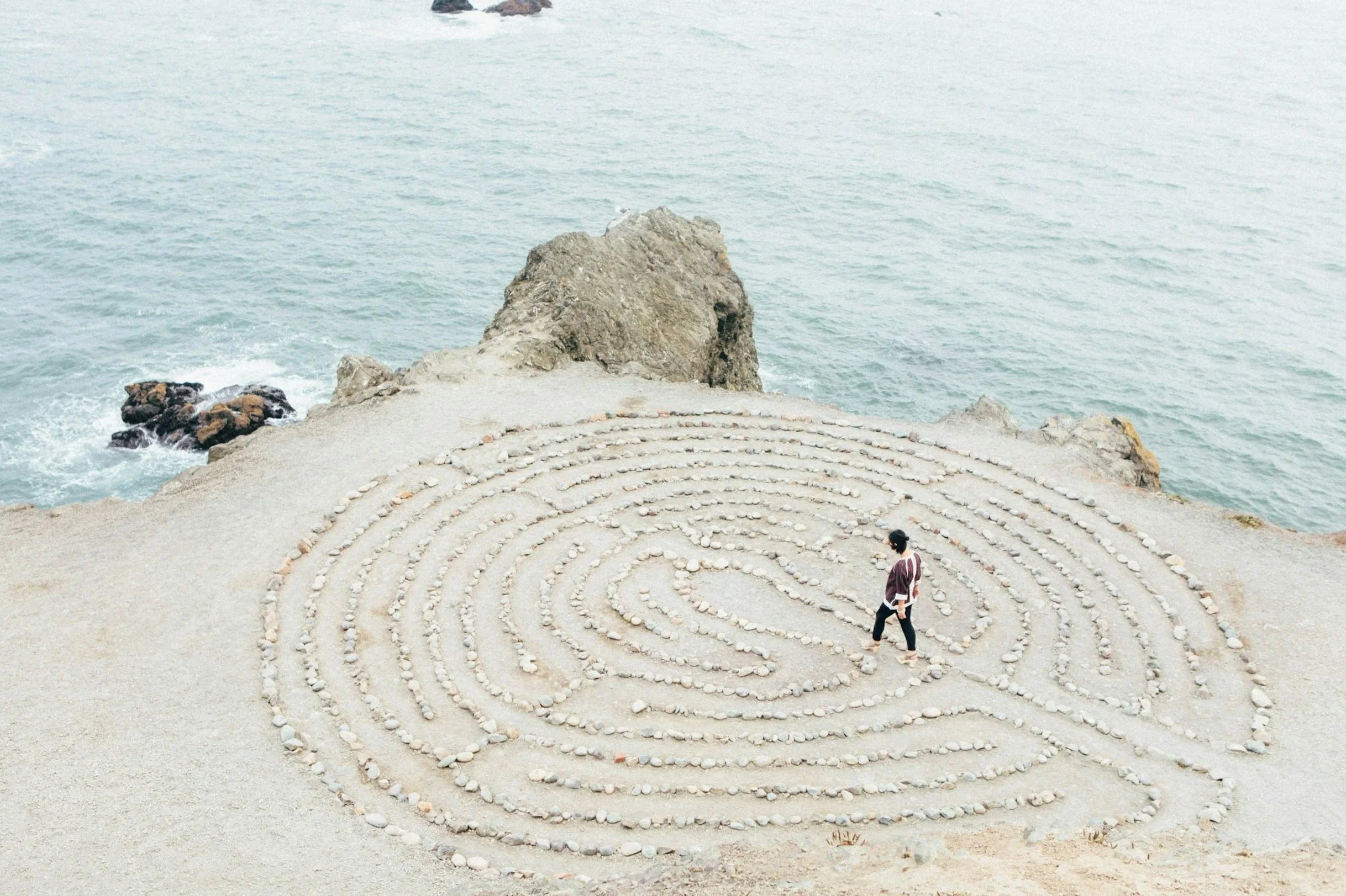 A person walking through a circular pebble maze on a sandy surface by the ocean with rocks and water in the background.