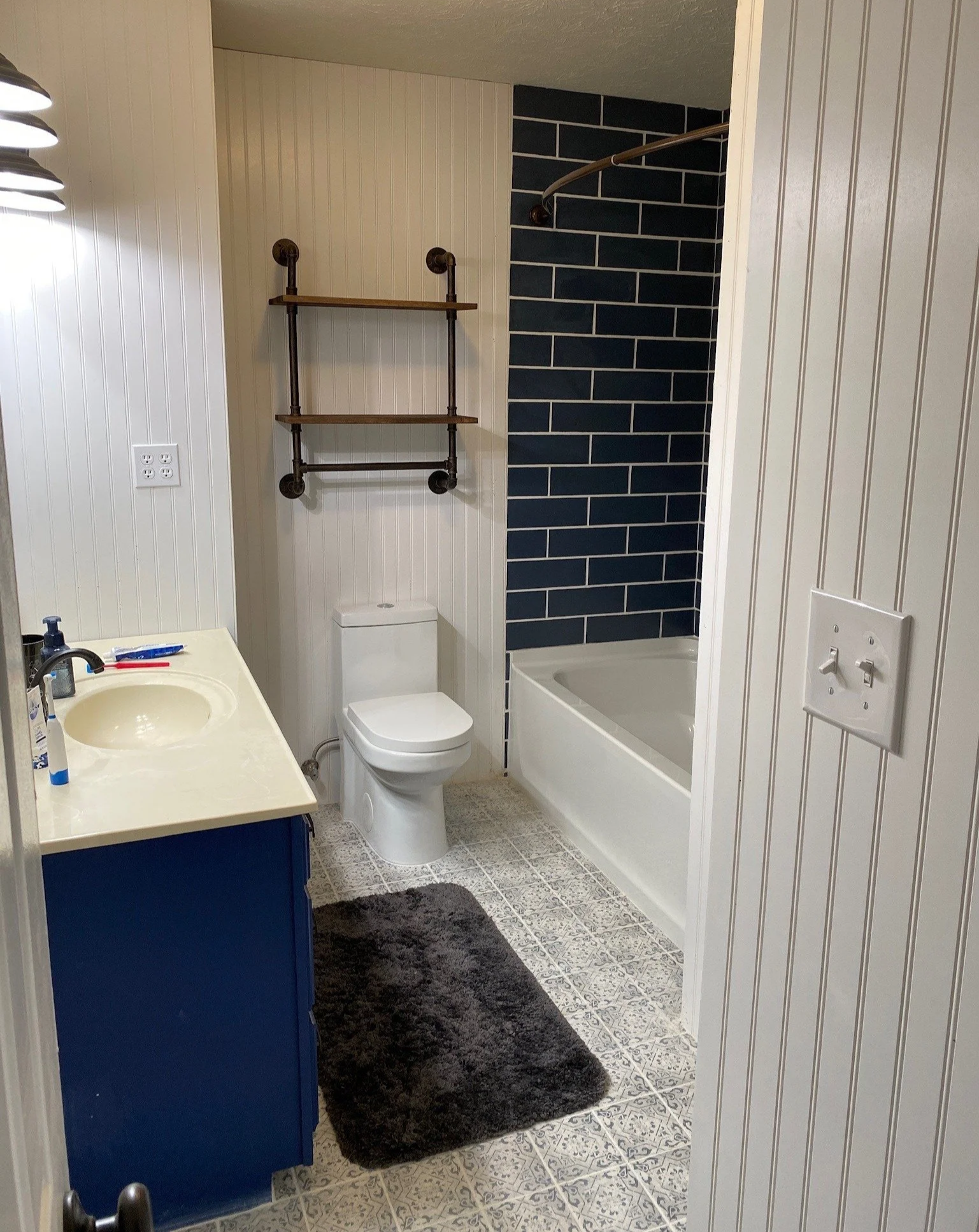 Bathroom with a white toilet, a vanity with a sink, a dark blue cabinet, a black rug, and a bathtub with a dark blue tiled wall behind it.