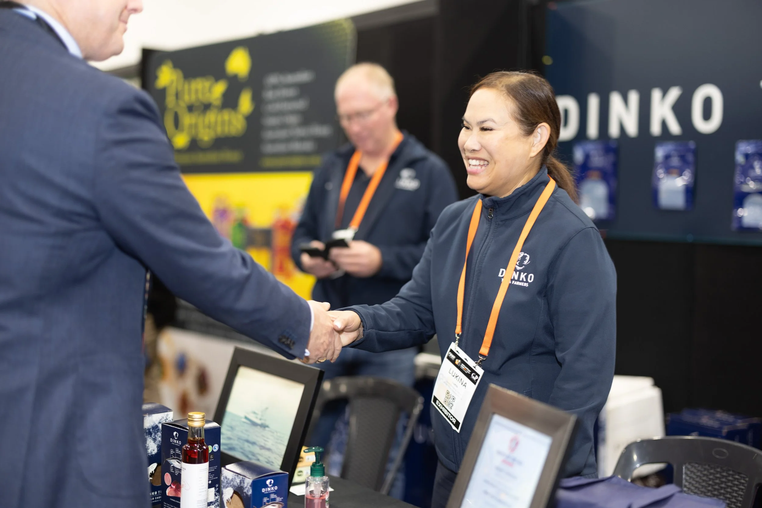 A woman in a navy jacket with an orange lanyard shaking hands with a man in a suit at a trade show booth. She is smiling. In the background, another woman is looking at her phone. The booth has a black and yellow display with the logo 'DINKO', products, and promotional materials.