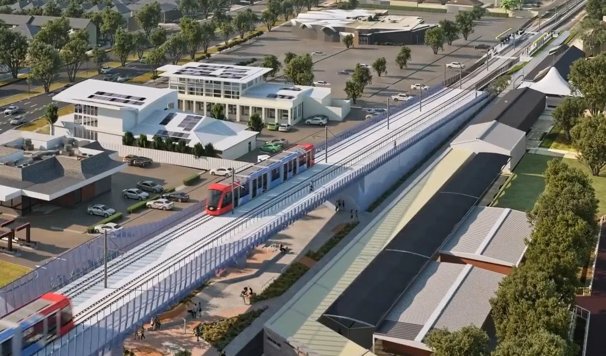 A modern urban transit station with a light rail train on elevated tracks, surrounded by commercial buildings, parking lots, and trees.