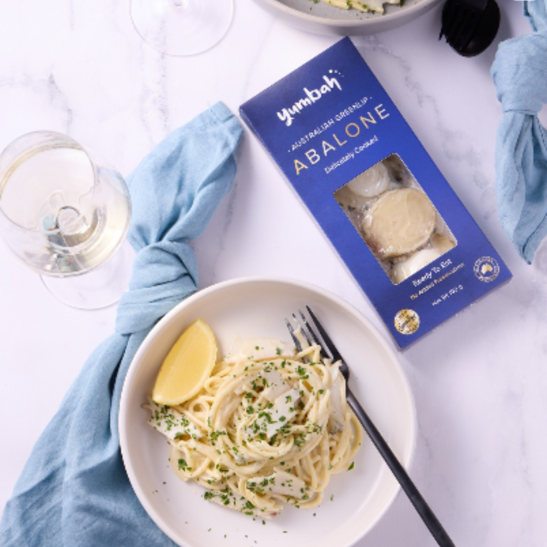 A white plate of spaghetti with lemon wedge and chopped herbs, a glass of white wine, a box of abalone, and a bowl of salad on a white marble table with blue cloth napkins.