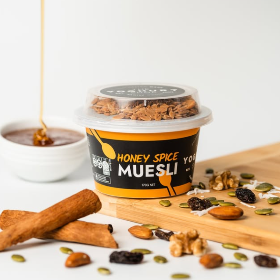 A tub of honey spice yogurt muesli with granola and mixed nuts, alongside cinnamon sticks and scattered pumpkin seeds and dried fruits on a wooden surface, with a bowl of honey being poured.