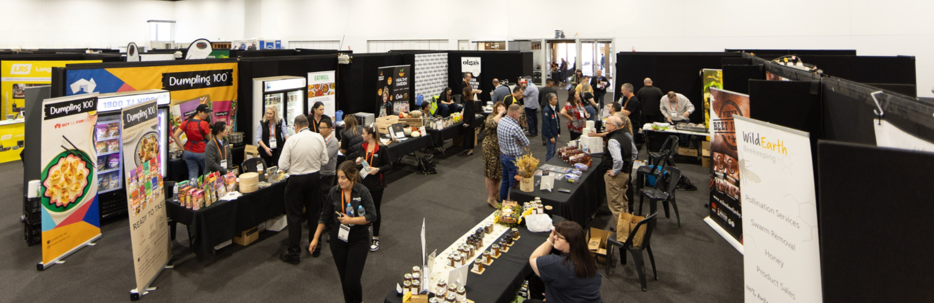 An indoor trade show or expo with multiple booths and attendees. People are browsing and talking at various vendor tables offering products and services, with displays and banners behind them.