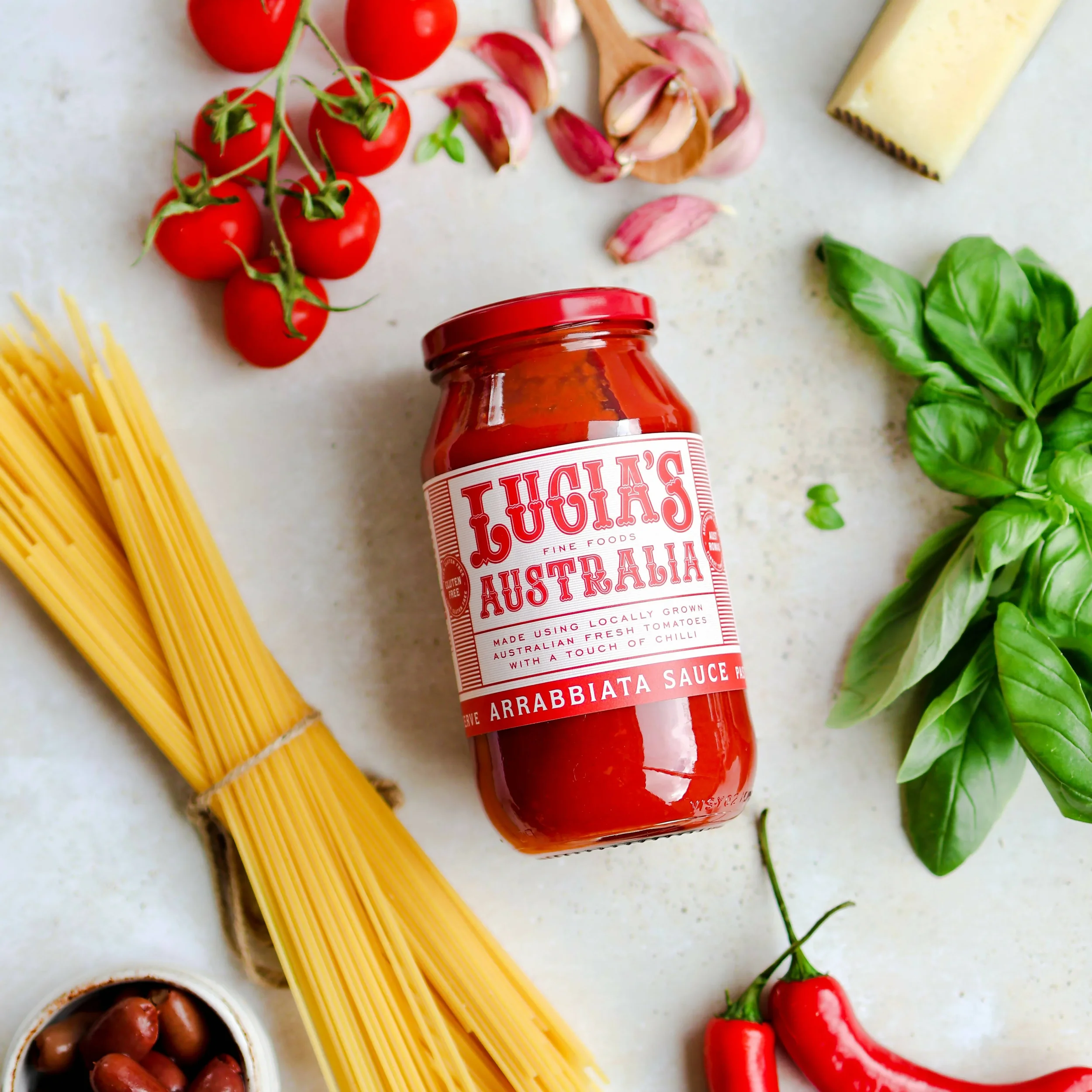 A jar of Luigi's Australia arrabbiata sauce surrounded by cherry tomatoes, garlic cloves, basil, uncooked spaghetti, olives, and red chili peppers on a white surface.