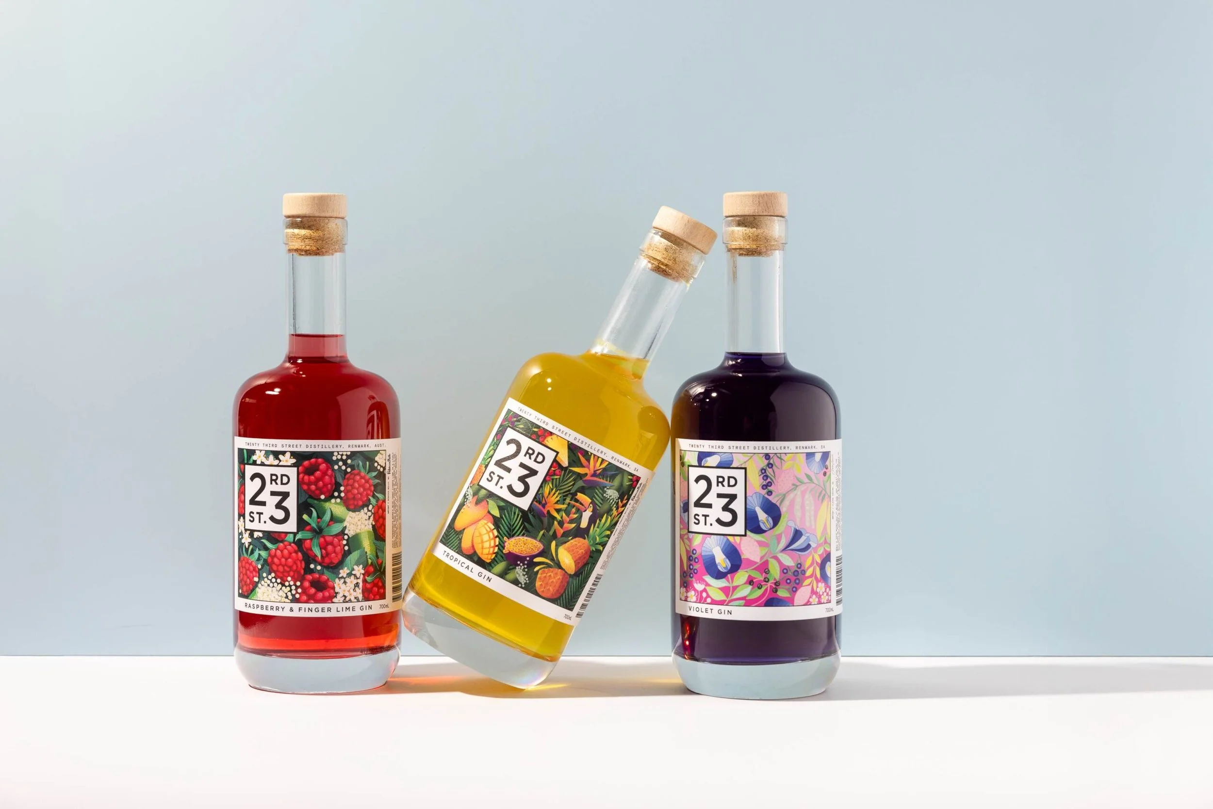 Three bottles of flavored gin with colorful labels, one tilted, on a white surface with a light blue background.