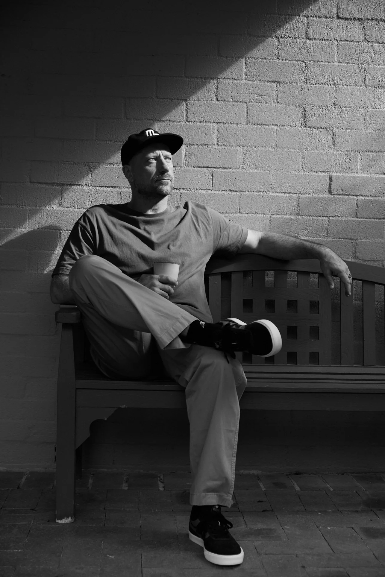 A man sitting on a wooden bench against a brick wall, holding a small cup, wearing a cap, t-shirt, and pants, with one arm resting on the bench's backrest, black and white photo.